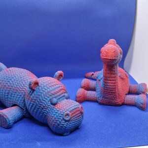 May include: Two 3D printed animal figurines, a blue and red hippopotamus and a blue and red dinosaur, on a blue background.