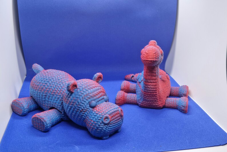 May include: Two 3D printed animal figurines, a blue and red hippopotamus and a blue and red dinosaur, on a blue background.