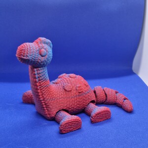 May include: A red and blue plastic dinosaur toy with articulated limbs. The dinosaur is lying on its belly with its head raised.