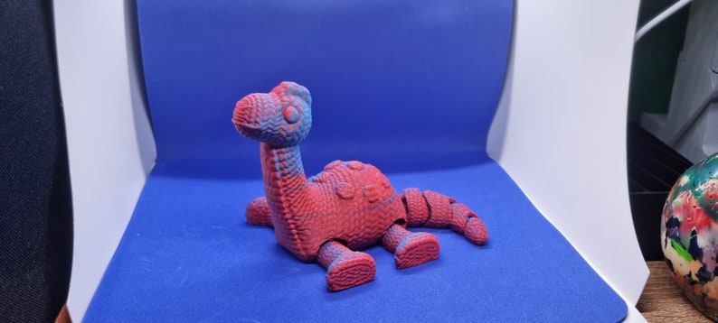 May include: A red and blue plastic dinosaur toy with articulated limbs. The dinosaur is lying on its belly with its head raised.
