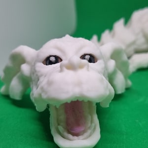Op de afbeelding: Witte 3D-geprinte drakenkop met een open mond en zwarte ogen. De drakenkop ligt op een groene ondergrond.