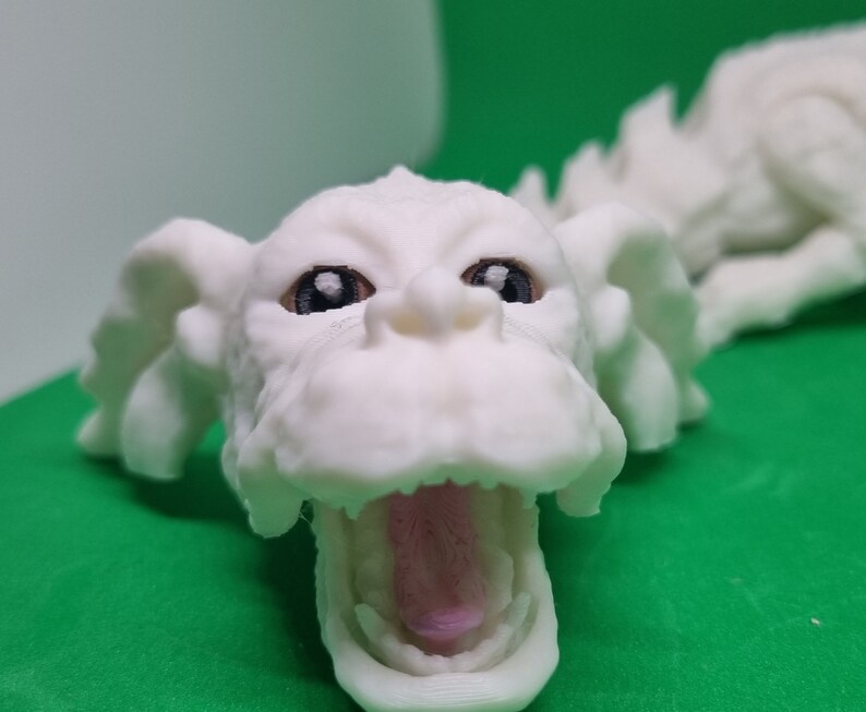 Op de afbeelding: Witte 3D-geprinte drakenkop met een open mond en zwarte ogen. De drakenkop ligt op een groene ondergrond.