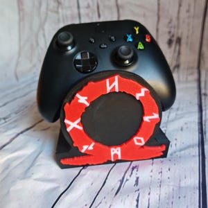 Peut inclure: Support de manette de jeu vid&eacute;o noir avec un logo God of War rouge et blanc. La manette est noire et a une disposition de boutons noir et blanc.