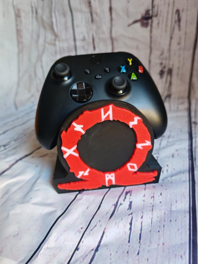 Peut inclure: Support de manette de jeu vid&eacute;o noir avec un logo God of War rouge et blanc. La manette est noire et a une disposition de boutons noir et blanc.