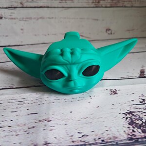 Op de afbeelding: Een tealgroen beeldje van een personage uit de Star Wars-franchise. Het beeldje heeft grote oren en een groot hoofd met zwarte ogen.