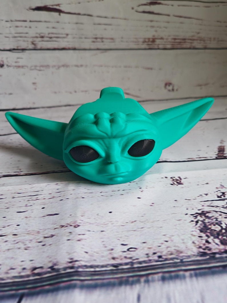 Op de afbeelding: Een tealgroen beeldje van een personage uit de Star Wars-franchise. Het beeldje heeft grote oren en een groot hoofd met zwarte ogen.
