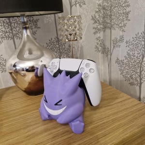 May include: A purple Gengar Pokemon figure holding a white Playstation 5 controller. The figure is smiling with a mischievous grin.