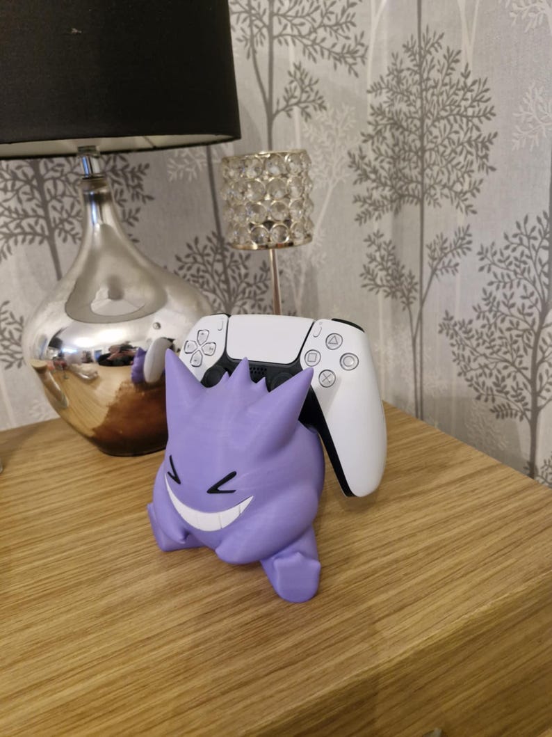 May include: A purple Gengar Pokemon figure holding a white Playstation 5 controller. The figure is smiling with a mischievous grin.