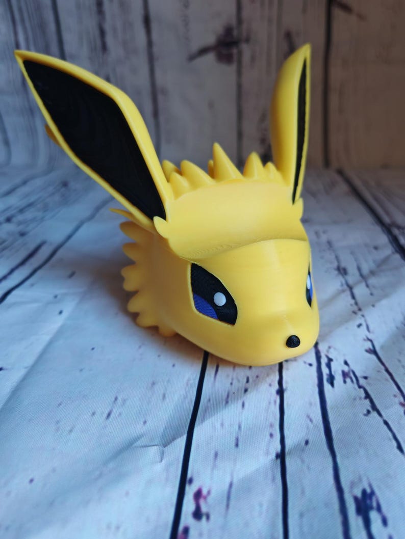 Peut inclure: Une figurine en 3D imprim&eacute;e en jaune d'un personnage Pokemon avec des oreilles noires et des yeux bleus. La figurine est un portrait du personnage.
