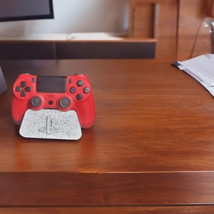 May include: A red PlayStation 4 controller is sitting on a white stand with the PlayStation logo on it. The controller is on a wooden table.