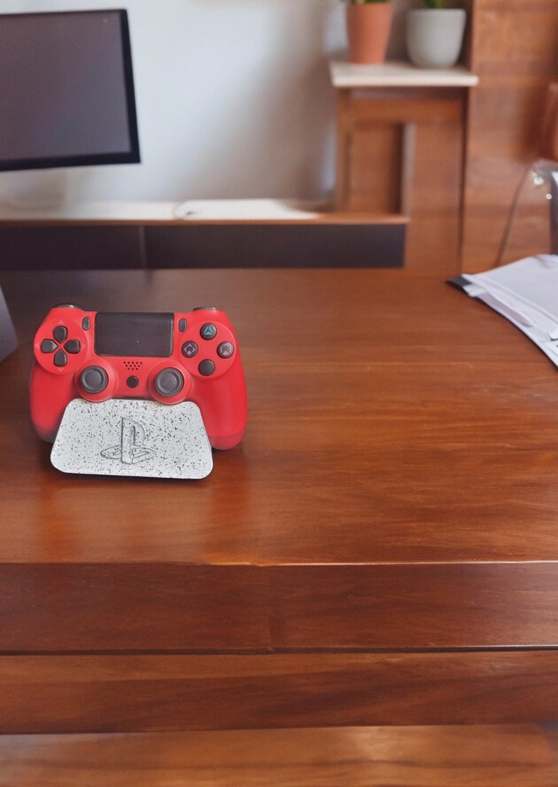 May include: A red PlayStation 4 controller is sitting on a white stand with the PlayStation logo on it. The controller is on a wooden table.