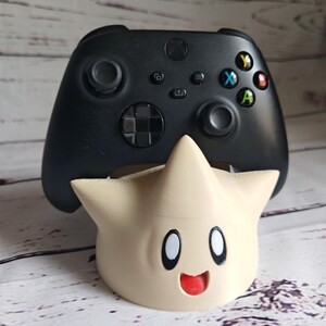 May include: A black Xbox controller sits on a beige 3D printed stand shaped like a star with a smiling face.