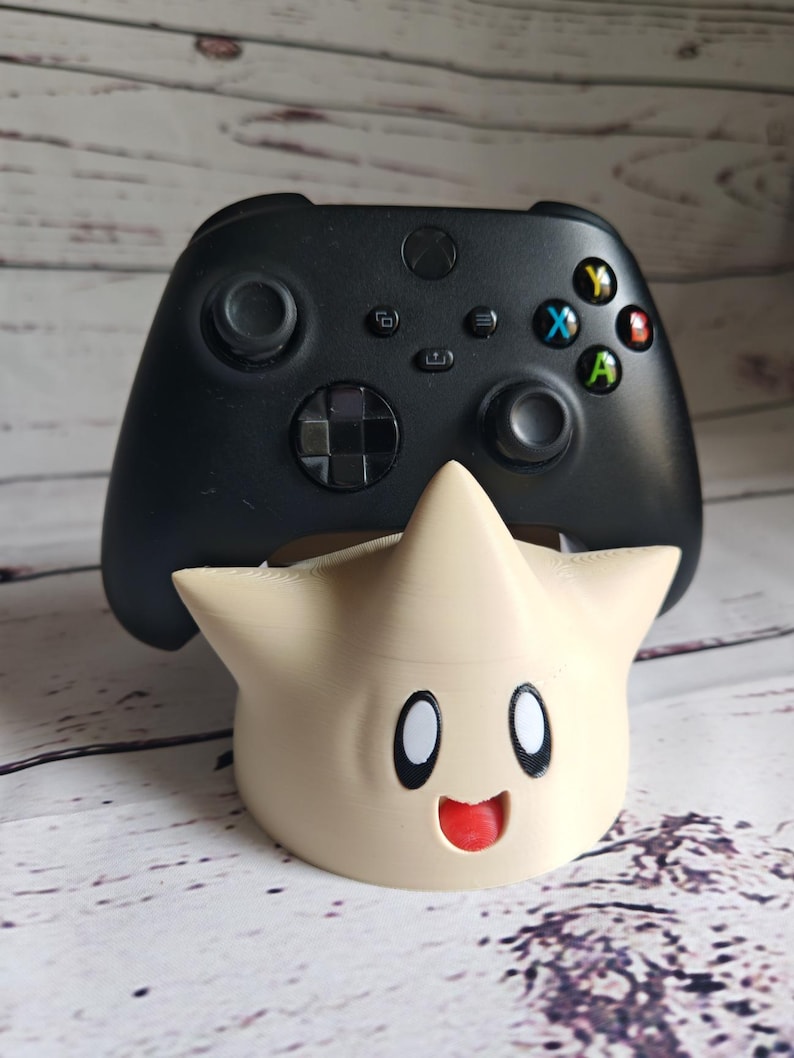 May include: A black Xbox controller sits on a beige 3D printed stand shaped like a star with a smiling face.