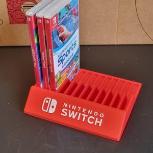 May include: Red plastic stand with slots for storing Nintendo Switch game cartridges. The stand has the Nintendo Switch logo and the words "Nintendo Switch" printed on the front. Three game cartridges are displayed in the stand, including "Nintendo Switch Sports", "Pok&eacute;mon Sword", and "Just Dance 2022".
