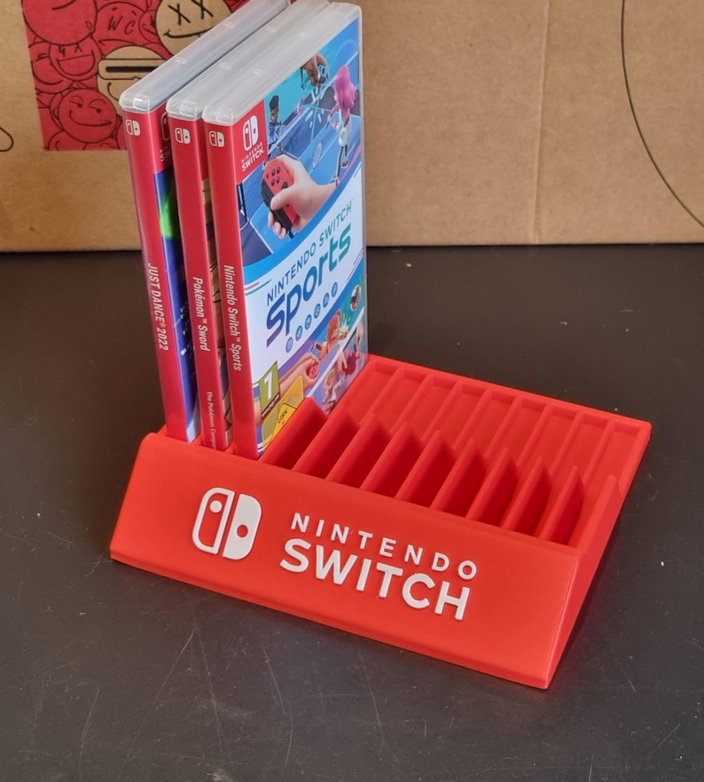 May include: Red plastic stand with slots for storing Nintendo Switch game cartridges. The stand has the Nintendo Switch logo and the words "Nintendo Switch" printed on the front. Three game cartridges are displayed in the stand, including "Nintendo Switch Sports", "Pok&eacute;mon Sword", and "Just Dance 2022".