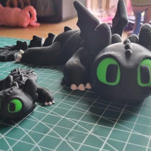 May include: Two black dragon figurines with green eyes. The larger dragon is lying down with its wings spread out. The smaller dragon is standing on its hind legs. Both dragons have a textured surface.