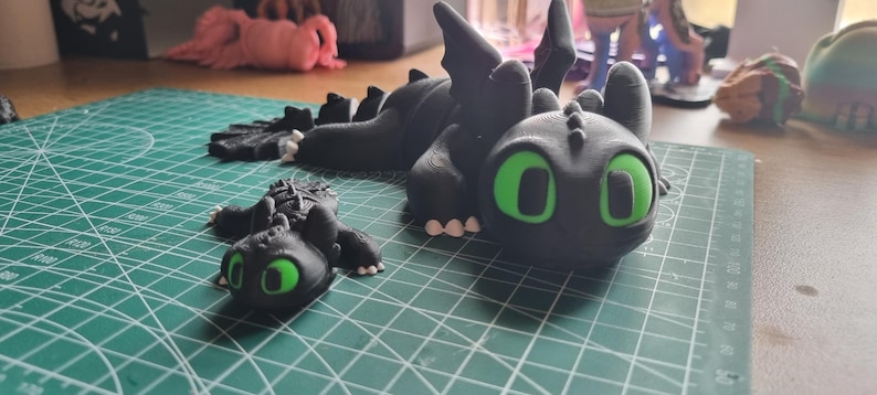 May include: Two black dragon figurines with green eyes. The larger dragon is lying down with its wings spread out. The smaller dragon is standing on its hind legs. Both dragons have a textured surface.