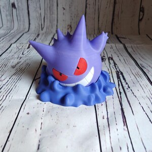 Może przedstawiać: Drukowana w 3D figurka w kolorze fioletowym i niebieskim przedstawiająca postać Pokemon, Gengar, z groźnym uśmiechem i czerwonymi oczami. Figurka jest ustawiona tak, jakby unosiła się w powietrzu.