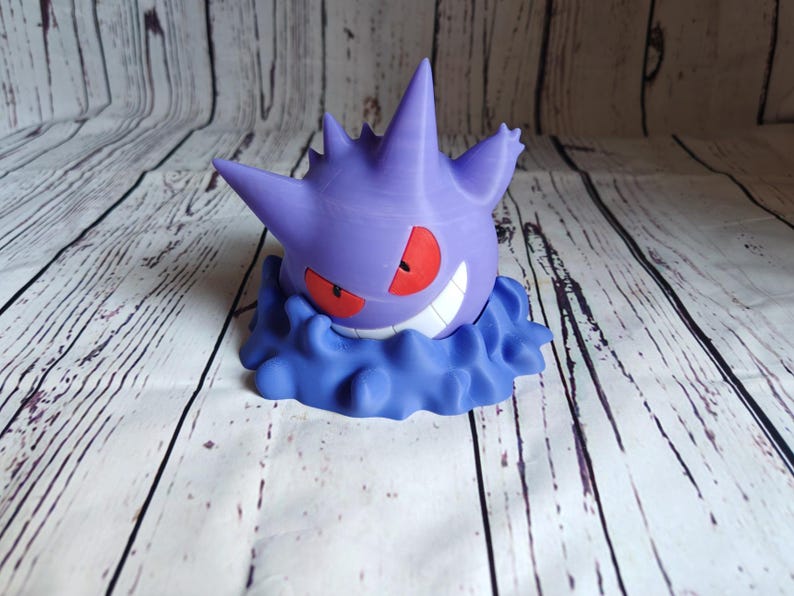 Może przedstawiać: Drukowana w 3D figurka w kolorze fioletowym i niebieskim przedstawiająca postać Pokemon, Gengar, z groźnym uśmiechem i czerwonymi oczami. Figurka jest ustawiona tak, jakby unosiła się w powietrzu.