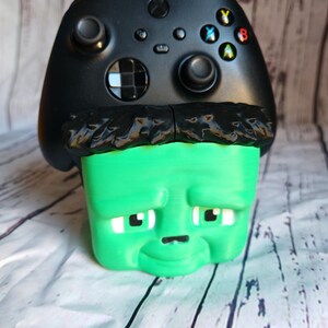 K&ouml;nnte beinhalten: Ein schwarzer Xbox-Controller liegt auf einer gr&uuml;nen, w&uuml;rfelf&ouml;rmigen Frankenstein-Figur. Die Figur hat schwarze Haare, wei&szlig;e Augen und eine kleine schwarze Nase. Der Controller hat farbige Tasten und zwei Joysticks.