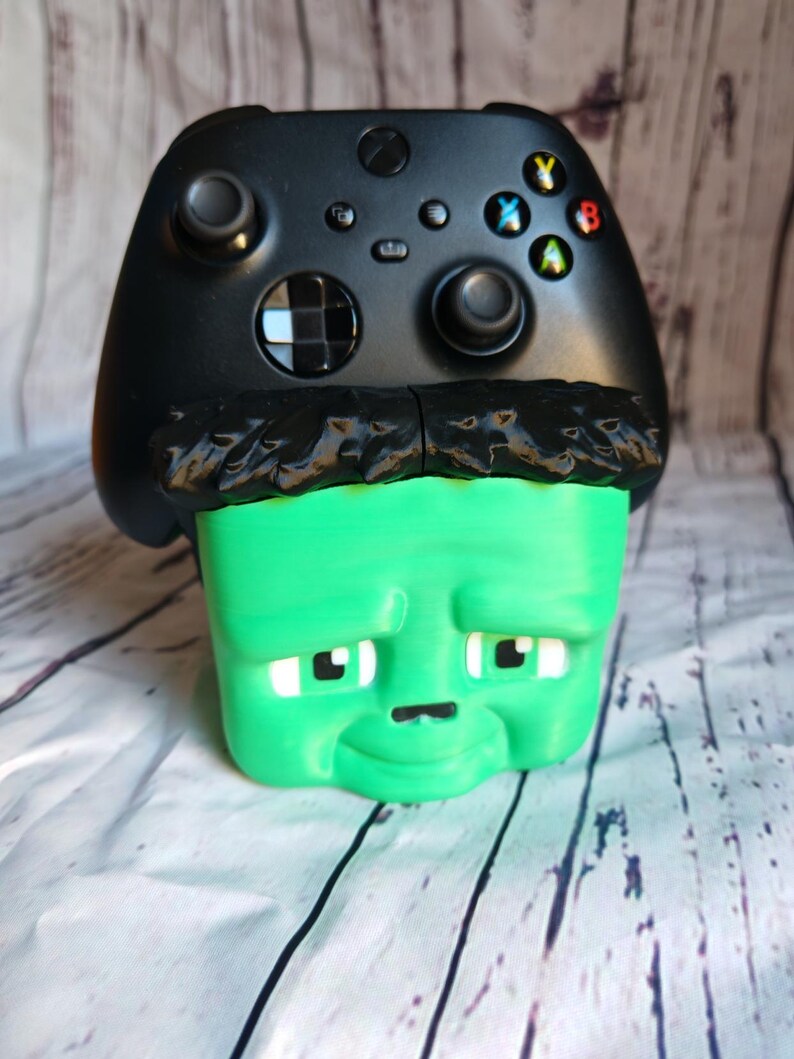 K&ouml;nnte beinhalten: Ein schwarzer Xbox-Controller liegt auf einer gr&uuml;nen, w&uuml;rfelf&ouml;rmigen Frankenstein-Figur. Die Figur hat schwarze Haare, wei&szlig;e Augen und eine kleine schwarze Nase. Der Controller hat farbige Tasten und zwei Joysticks.