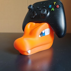 Op de afbeelding: Oranje en blauwe Pokemon-figuur met een zwarte Xbox-controller die op zijn hoofd rust.