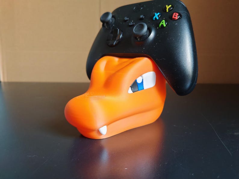 Op de afbeelding: Oranje en blauwe Pokemon-figuur met een zwarte Xbox-controller die op zijn hoofd rust.