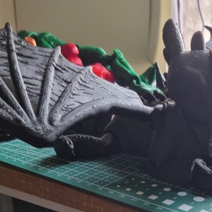 K&ouml;nnte beinhalten: Schwarzes Drachenspielzeug mit gro&szlig;en Fl&uuml;geln, das auf einer gr&uuml;nen Schneidematte liegt. Der Drache hat einen strukturierten, schuppigen K&ouml;rper und ein detailliertes Gesicht.