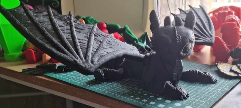 K&ouml;nnte beinhalten: Schwarzes Drachenspielzeug mit gro&szlig;en Fl&uuml;geln, das auf einer gr&uuml;nen Schneidematte liegt. Der Drache hat einen strukturierten, schuppigen K&ouml;rper und ein detailliertes Gesicht.