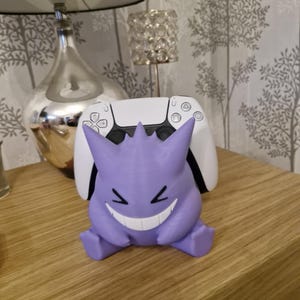 May include: A purple and white 3D printed Ghost Pokemon figure holding a white Playstation 5 controller. The figure is sitting on a wooden surface.