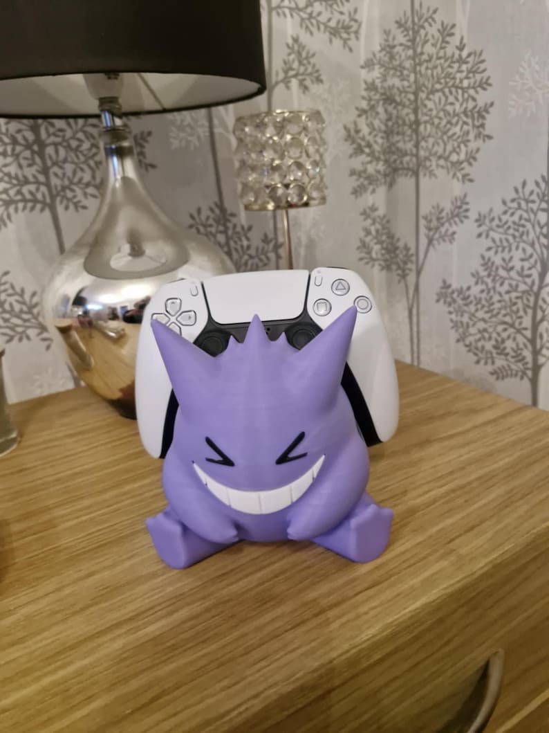 May include: A purple and white 3D printed Ghost Pokemon figure holding a white Playstation 5 controller. The figure is sitting on a wooden surface.