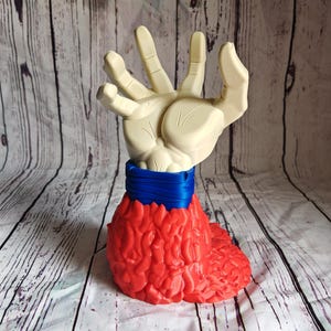 Pode incluir: Uma escultura &uacute;nica com uma m&atilde;o creme emergindo de uma base vermelha texturizada. Uma faixa azul envolve a &aacute;rea do pulso da m&atilde;o. A escultura est&aacute; em um fundo com um padr&atilde;o de gr&atilde;o de madeira clara.