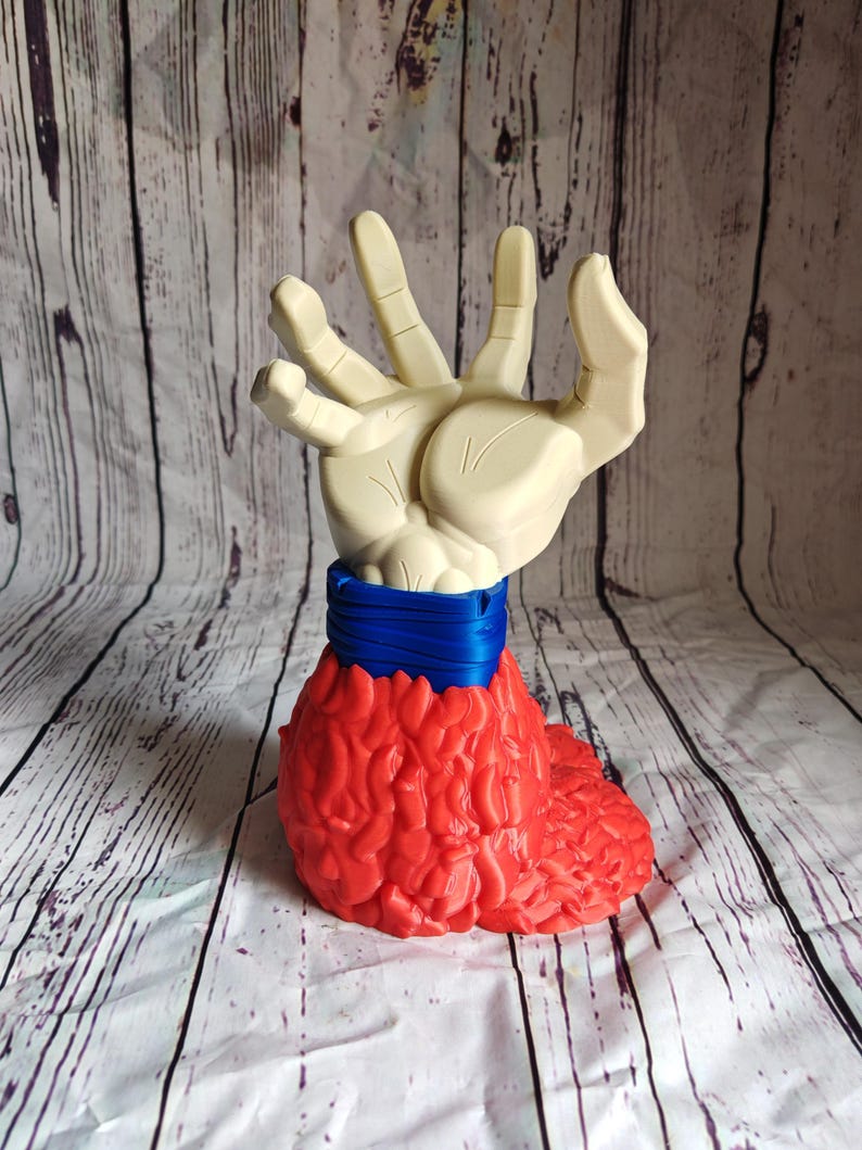 Pode incluir: Uma escultura &uacute;nica com uma m&atilde;o creme emergindo de uma base vermelha texturizada. Uma faixa azul envolve a &aacute;rea do pulso da m&atilde;o. A escultura est&aacute; em um fundo com um padr&atilde;o de gr&atilde;o de madeira clara.
