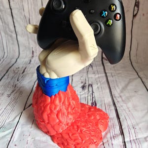 Pode incluir: Um controle Xbox preto segurado por um suporte em forma de m&atilde;o creme. O suporte &eacute; preso a uma base vermelha texturizada com um detalhe azul. O controle tem bot&otilde;es coloridos e um direcional.