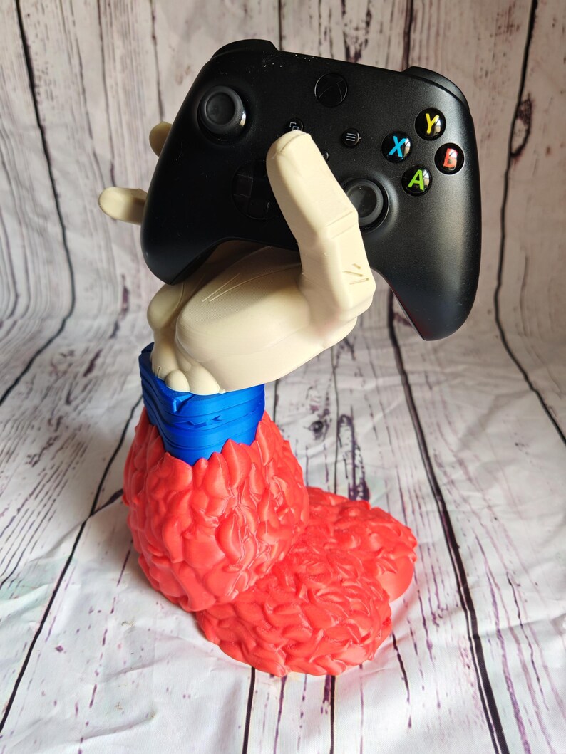 Pode incluir: Um controle Xbox preto segurado por um suporte em forma de m&atilde;o creme. O suporte &eacute; preso a uma base vermelha texturizada com um detalhe azul. O controle tem bot&otilde;es coloridos e um direcional.