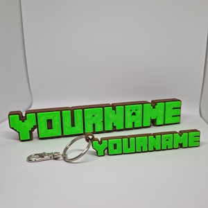 Peut inclure: Un porte-cl&eacute;s Minecraft en brun et vert avec le mot "YOURNAME" en lettres pixelis&eacute;es. Le porte-cl&eacute;s est attach&eacute; &agrave; un anneau de cl&eacute; argent&eacute;.