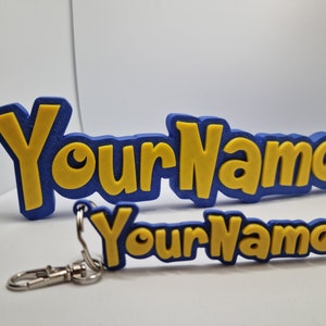 K&ouml;nnte beinhalten: Ein blau-gelbes Namensschild und Schl&uuml;sselanh&auml;nger. Der Name "YourName" ist gelb dargestellt, blau umrandet. Der Schl&uuml;sselanh&auml;nger hat einen silbernen Verschluss. Das Schild ist 3D-gedruckt.