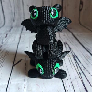 May include: A stack of two black knitted dragon figurines with green eyes. The dragons are facing forward with their wings spread out.