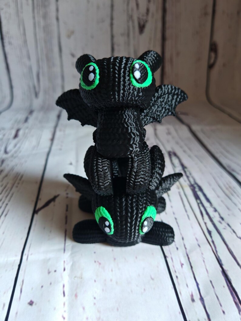 May include: A stack of two black knitted dragon figurines with green eyes. The dragons are facing forward with their wings spread out.