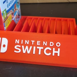 May include: Orange plastic Nintendo Switch game case holder with slots for 12 game cartridges. The holder has the Nintendo Switch logo and text on the front.