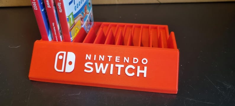 May include: Orange plastic Nintendo Switch game case holder with slots for 12 game cartridges. The holder has the Nintendo Switch logo and text on the front.