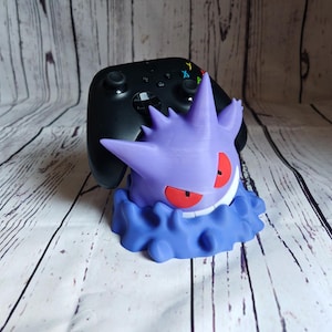 Może przedstawiać: Fioletowa i niebieska postać Pokemon Gengar trzymająca czarny kontroler Xbox. Kontroler spoczywa na głowie Gengara. Gengar stoi na białym drewnianym podłożu.