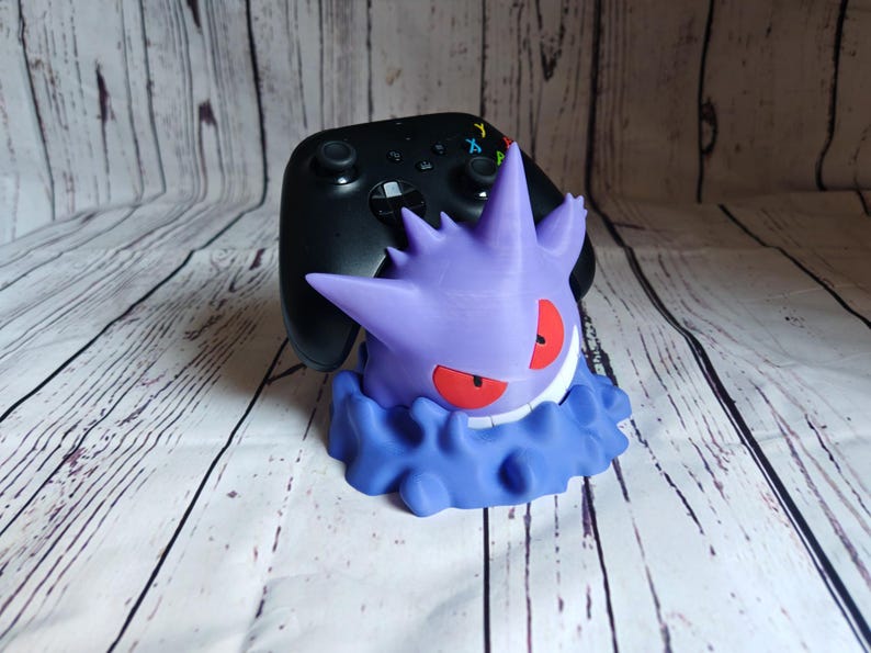 Może przedstawiać: Fioletowa i niebieska postać Pokemon Gengar trzymająca czarny kontroler Xbox. Kontroler spoczywa na głowie Gengara. Gengar stoi na białym drewnianym podłożu.