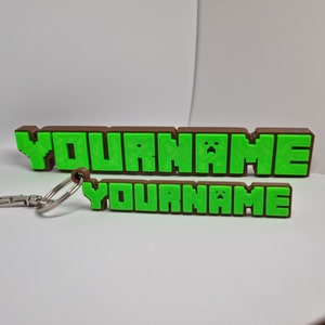 Peut inclure: Un porte-cl&eacute;s brun et vert avec le texte "YOURNAME" dans une police de caract&egrave;res pix&eacute;lis&eacute;e. Le porte-cl&eacute;s est en plastique et poss&egrave;de un anneau en m&eacute;tal.