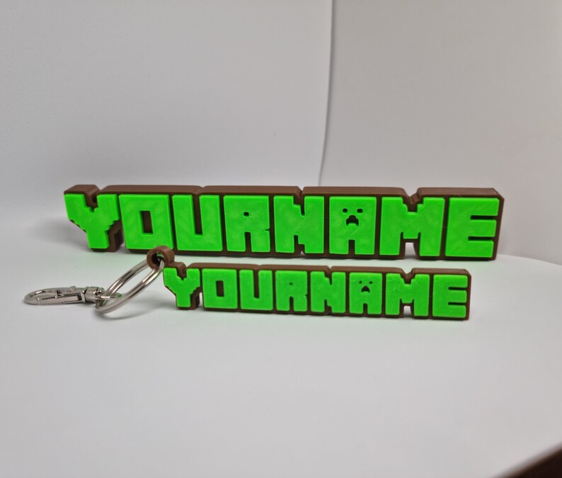 Peut inclure: Un porte-cl&eacute;s brun et vert avec le texte "YOURNAME" dans une police de caract&egrave;res pix&eacute;lis&eacute;e. Le porte-cl&eacute;s est en plastique et poss&egrave;de un anneau en m&eacute;tal.