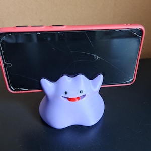 以下が含まれることがあります： ポケモンのキャラクター、ゲンガーの形をした、紫と黒の3Dプリントされた携帯電話スタンド。スタンドは、ひび割れた画面のピンクの携帯電話を持っています。