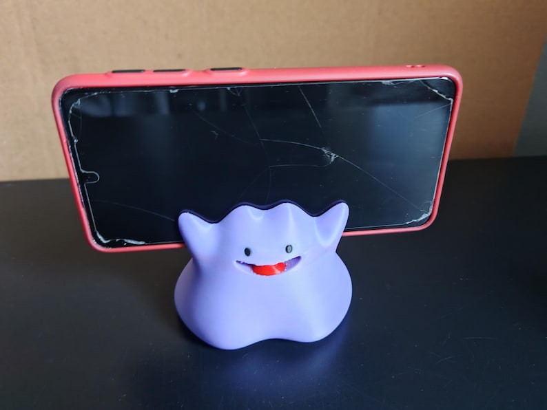 以下が含まれることがあります： ポケモンのキャラクター、ゲンガーの形をした、紫と黒の3Dプリントされた携帯電話スタンド。スタンドは、ひび割れた画面のピンクの携帯電話を持っています。