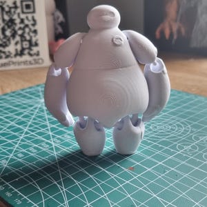 May include: White 3D printed figurine of a cartoon character with a round body and short arms and legs. The character has a large head with a simple face and a small mouth. The figurine is standing on a green cutting mat.