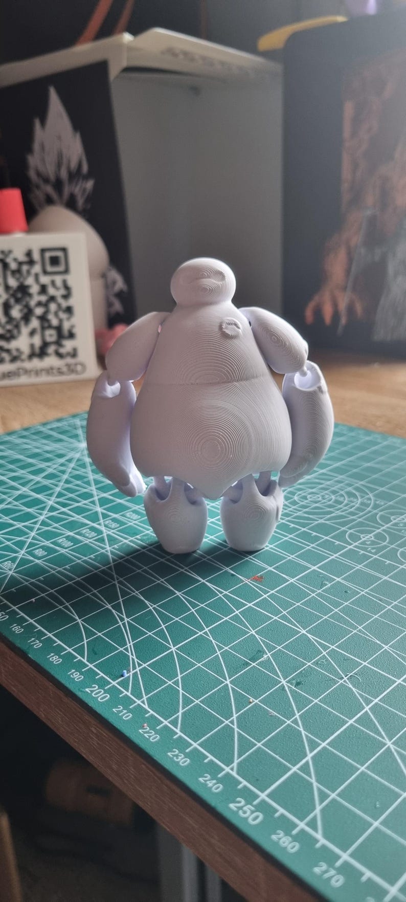 May include: White 3D printed figurine of a cartoon character with a round body and short arms and legs. The character has a large head with a simple face and a small mouth. The figurine is standing on a green cutting mat.