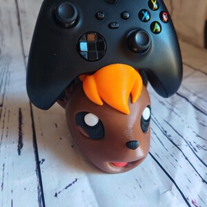 Pode incluir: Um controlador Xbox preto est&aacute; em cima da cabe&ccedil;a de um personagem Pokemon impresso em 3D de cor marrom e laranja.